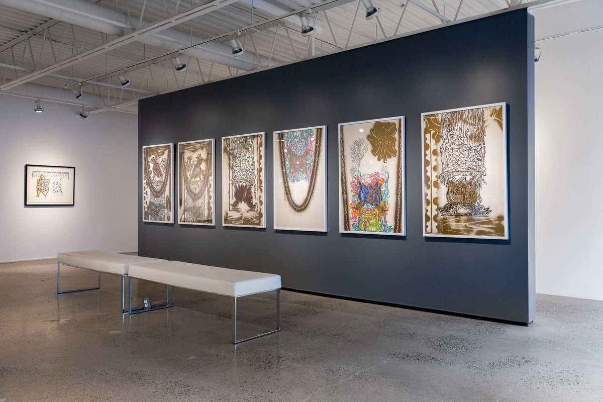 A gallery interior showing a dark blue wall with six framed abstract artworks in varying colors, alongside a white bench on a polished concrete floor.