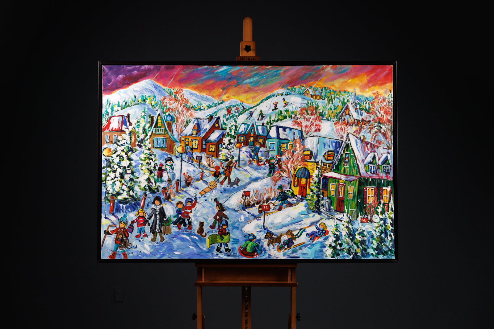 Colorful winter village scene painting on an easel against a black background