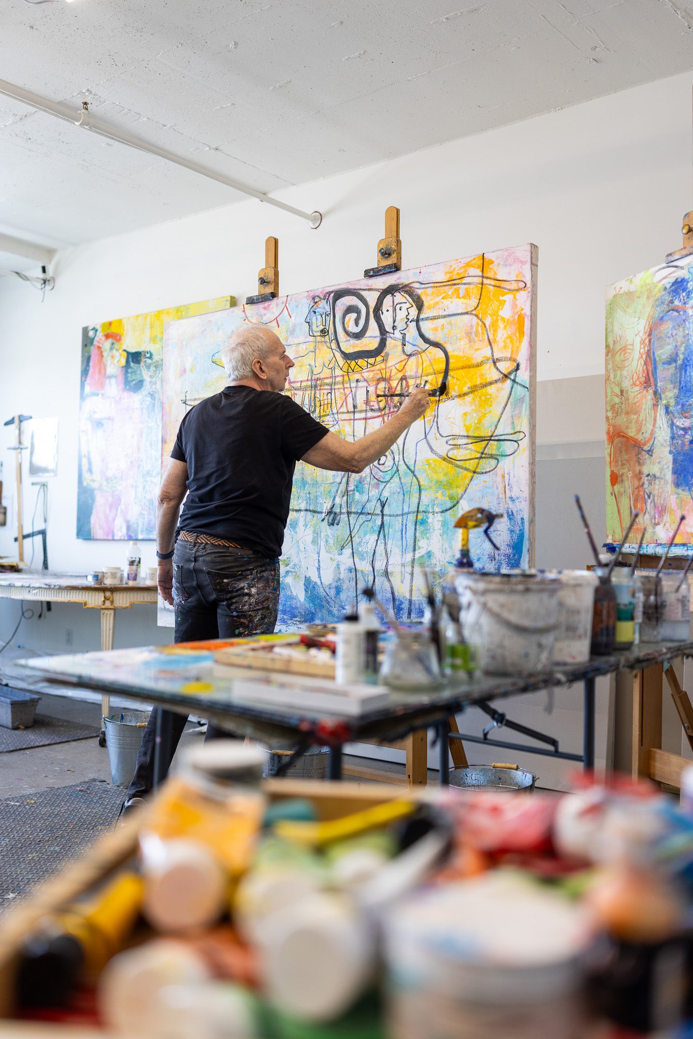 Artist painting a large abstract canvas in a studio with various art supplies on a table.