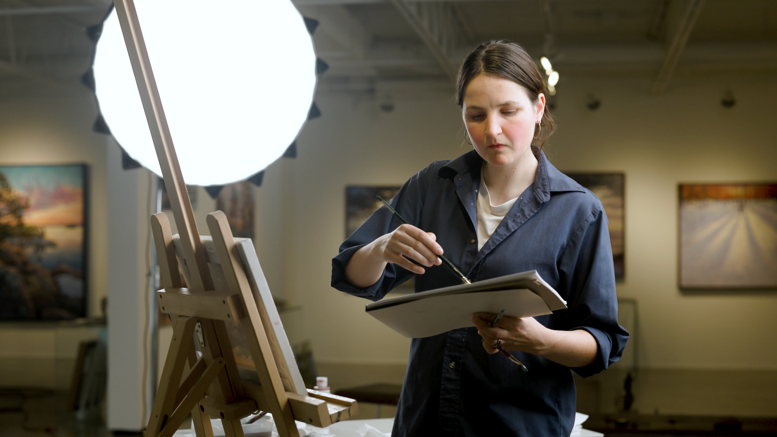 Woman in an art studio holding a tablet next to an easel with a painting.